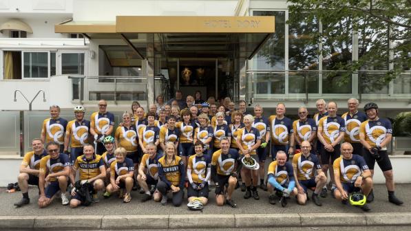 A big mixed group of people in matching cycling kit are posing outside a building