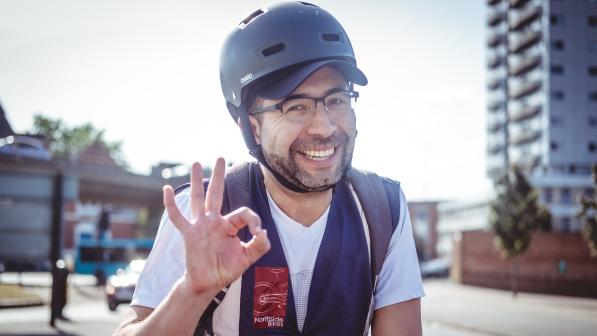A smiling man wearing a helmet, glasses, and a cycling vest gives an ‘OK’ hand gesture while out on a sunny city street, with buildings and traffic in the background.