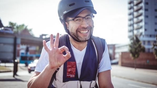 A smiling man wearing a helmet, glasses, and a cycling vest gives an ‘OK’ hand gesture while out on a sunny city street, with buildings and traffic in the background.