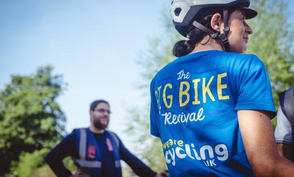 A woman wearing a blue T-shirt and cycle helmet is standing with her back to the camera. The T-shirt reads 'The Big Bike Revival; We are Cycling UK'
