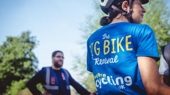 A woman wearing a blue T-shirt and cycle helmet is standing with her back to the camera. The T-shirt reads 'The Big Bike Revival; We are Cycling UK'