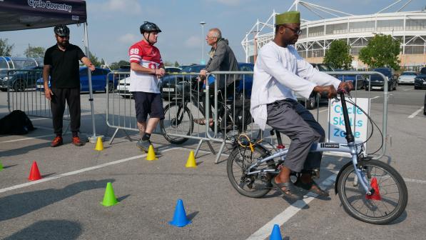 A man in a turban, tunic and trousers is riding a folding bike around an obstacle course. There are two men in the background watching him and another on a bike waiting to have a go
