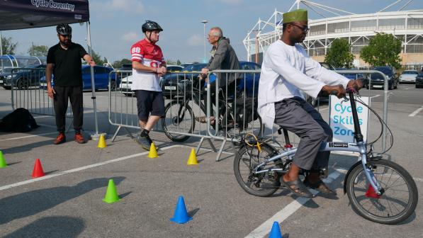 A man wearing traditional clothing and a green cap rides a small folding bike through a course marked with coloured cones in a Cycle Skills Zone in a car park. Three other men wearing helmets and casual clothing stand nearby behind a metal barrier, observing. A Cycle Coventry tent and a stadium are visible in the background.