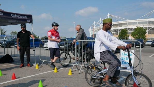 A man wearing traditional clothing and a green cap rides a small folding bike through a course marked with coloured cones in a Cycle Skills Zone in a car park. Three other men wearing helmets and casual clothing stand nearby behind a metal barrier, observing. A Cycle Coventry tent and a stadium are visible in the background.