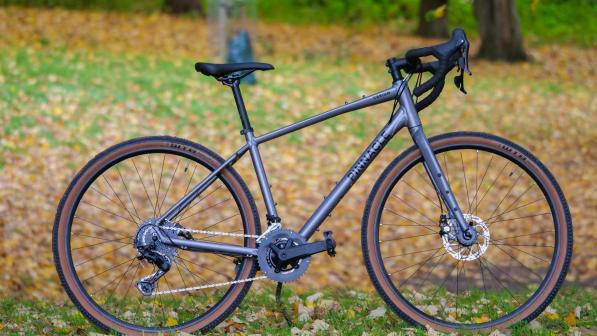 The Pinnacle Cerium, a silver grey gravel bike, propped up in a leaf-strewn field with trees in the background