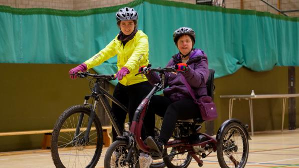Two people are on cycles in a sports hall. One is on a trike and wearing purple, the other is on a mountain bike and wearing yellow hi-vis