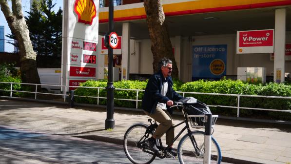 A man on a Dutch-style bike with a basket on the front is riding on a cycle path past a petrol station. He's wearing normal clothes, sunglasses and there's a bag in the basket