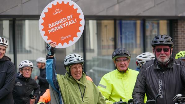 A group of people with bikes and in cycle helmets and hi-vis waterproofs are gathered together with one holding a sign reading 'Banned by Merseyrail' with the Cycling UK logo on it