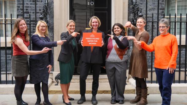 Seven women stand in front of the door of 10 Downing Street. They are smiling and pointing towards Cycling UK CEO Sarah Mitchell in the centre, who holds an orange box with the words 'Make cycling safer for women' on it