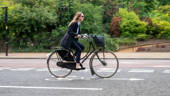A woman wearing normal clothes is riding a black Dutch-style bike on an urban road. There is a bag in the basket on the front. The bike has a chain guard and skirt guard