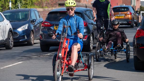 Person wearing a yellow helmet rides a red handcycle on a residential road between parked cars, with two other cyclists riding behind.