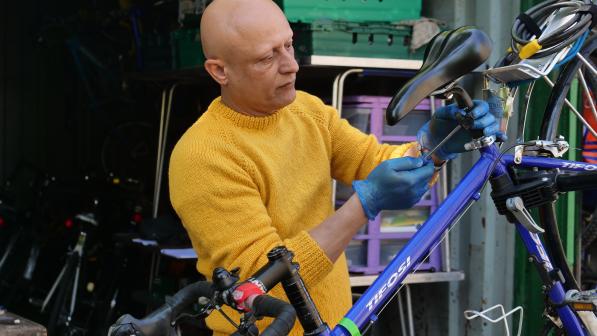 A man wearing a yellow jumper and blue gloves adjusts the rear brake of a blue Tifosi bicycle in a workshop setting, surrounded by bike tools and parts.