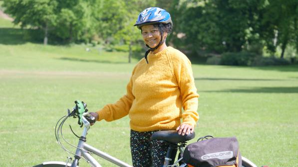 A woman wearing a blue cycle helmet and yellow jumper smiles while standing beside her bicycle in a grassy park. Her bike is equipped with panniers, and the background is filled with green trees.