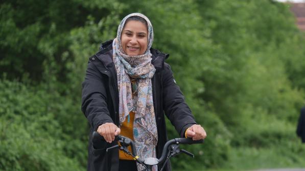 A woman wearing a patterned headscarf, black coat, and yellow jumper smiles while holding the handlebars of a bicycle on a tree-lined path.