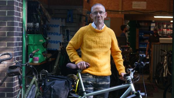 A man wearing a yellow jumper stands proudly beside a silver bicycle outside a bike workshop, with shelves and tools visible in the background.