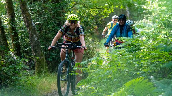 A group of people is riding on mountain bikes on an off-road track through a forest. They are wearing cycling kit and helmets and are smiling