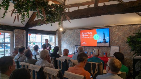 A man is standing at the front of a room with a big television in front him presenting to a room full of people