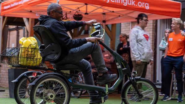 A man wearing a hoody and jeans is riding a trike past a gazebo with bright orange awning with the Cycling UK web address printed on it. There are lots of people standing around under the gazebo chatting
