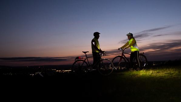 Two people stand with bikes on a hillside at dusk, almost silhouetted against the pink and grey sky