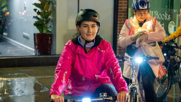 A young women wearing a bright pink jacket sits on her bike, on a dark evening in a town square