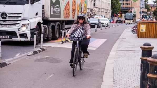 A woman in normal clothes with a shoulder bag and cycle helmet is riding a hybrid bike along a protected cycle path with traffic behind her, including a big lorry she's separated from