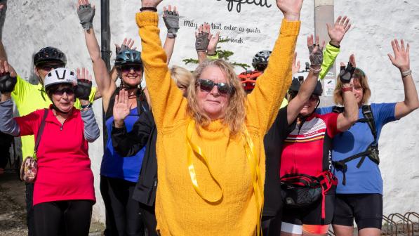 A woman wearing a yellow knitted sweater is standing with her hands up in a motion of celebration. She's in front of a group of people wearing cycle kit also holding their hands up. Behind them, on a the white outside wall of a building, is a sign that reads 'Bon Appetit'