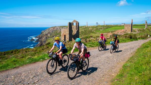 Cyclists riding along a coastal gravel trail.  There are ruins and the sea in the background.