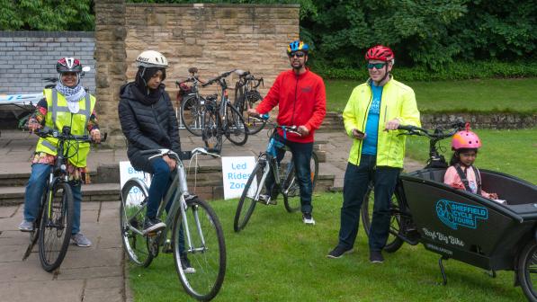 Group of adults and a child on bicycles preparing for a ride in a park. One child sits in a cargo bike marked 'Sheffield Cycle Tours'. Signs in the background read 'Led Rides Zone'. All riders are wearing helmets and outdoor clothing.