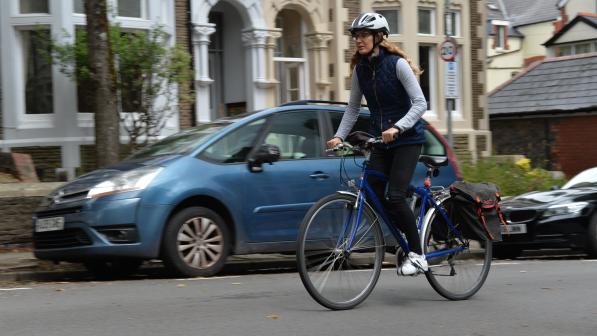 A woman on a blue hybrid bike, wearing normal clothes and a cycle helmet, is cycling past a parked car in an urban street