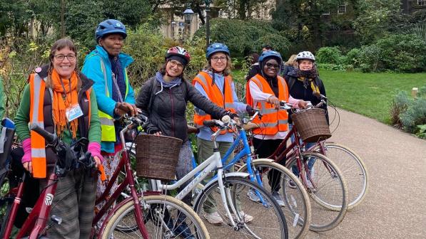 A group of women in a mix of clothing is standing with their bikes on a paved path in a park