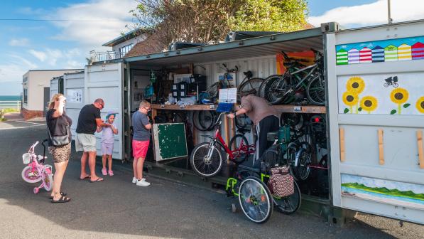 A group of people, including adults and children, gather around an open shipping container filled with bikes and cycling equipment. One person is retrieving a red bike, while others wait nearby. The container is painted with sunflowers and beach hut designs, and the seaside is visible in the background.