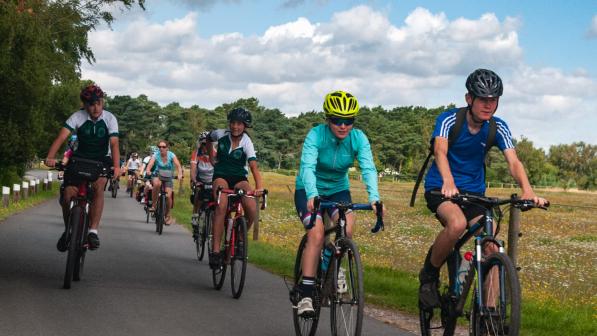 A large group of people is cycling along a singletrack road with a field to one side and trees to the other