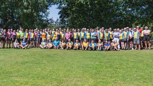 A large group of people are all gathered together on some grass with trees in the background. They are all wearing cycling clothing