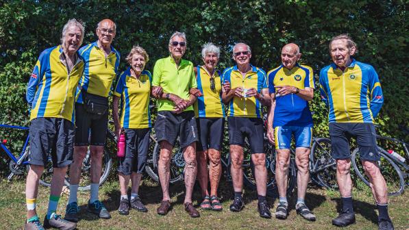A group of older men and women in cycling kit are standing in a line. There are bikes leaning against trees behind them