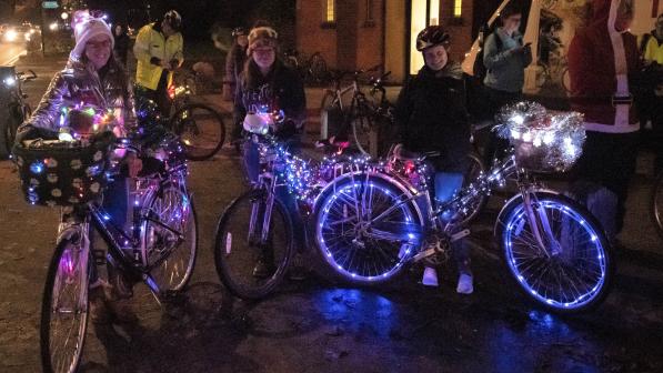 Three women stand smiling at the camera on a dark street, holding bikes decorated with fairy lights