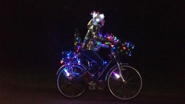 A female cyclist is riding a hybrid bike at night. It has a front basket and rear rack and there are loads of coloured lights all over it. She is wearing jeans, a gold jacket and a festive hat