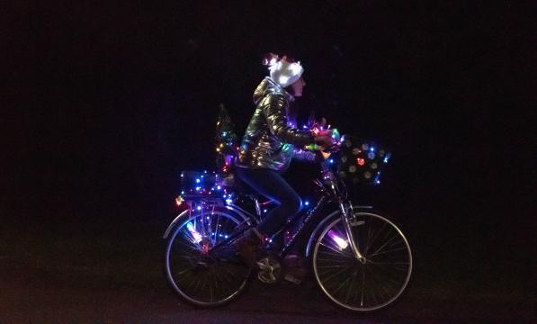 A female cyclist is riding a hybrid bike at night. It has a front basket and rear rack and there are loads of coloured lights all over it. She is wearing jeans, a gold jacket and a festive hat