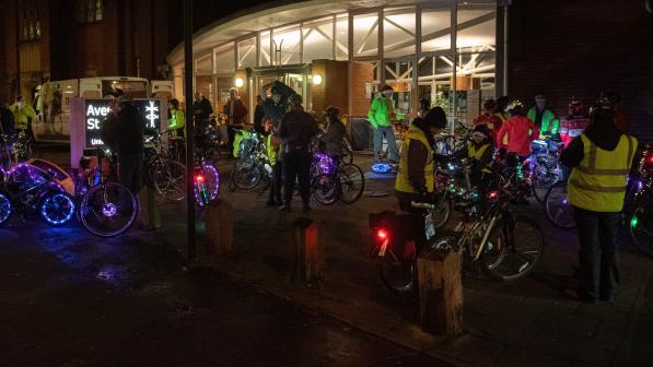 A group of cyclists gather outside a train station on a dark evening. Their bikes are lit with colourful fairy lights