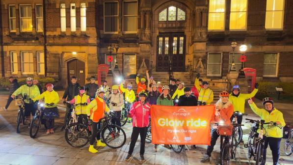 A big group of people are standing outside a town hall in the evening. They have bikes and are wearing cycling kit. They're holding a banner with the Cycling UK logo and the words 'Glow ride'
