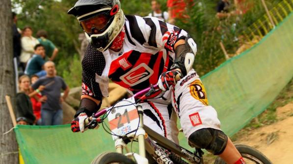 A woman is taking part in a mountain bike race. She is wearing a full-face helmet and goggles, and mountain biking clothing including knee and elbow pads. Her race number is 21 and she’s racing for Great Britain 