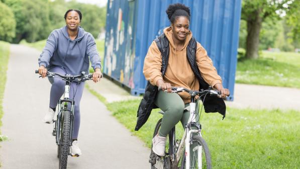 Two women are cycling in a park with a blue container behind them. They're both wearing normal clothes and on hybrids