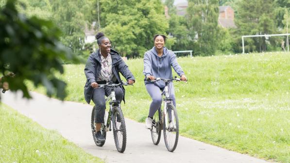 Two women are cycling on a paved path in a park. They are both riding hybrid bikes. One is wearing black jeans, trainers, a patterned shirt and grey coat. The other is wearing a grey tracksuit and white trainers. 