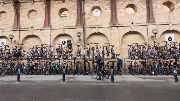 A person is wheeling a bike past several cycle racks with lots of bikes in them at a railway station