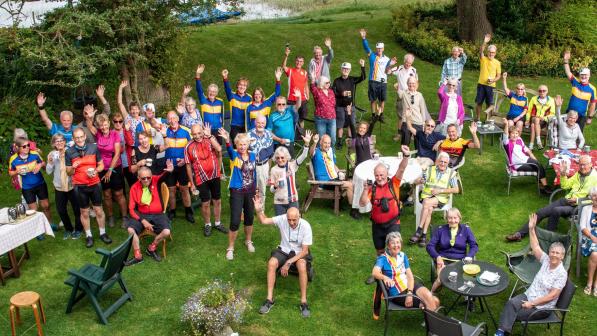 A big group of people, some sitting at tables, some standing, are in a field. Many are wearing cycling kit. There are plates and cups on the tables. Some of the people have their arms in the air in a celebratory gesture