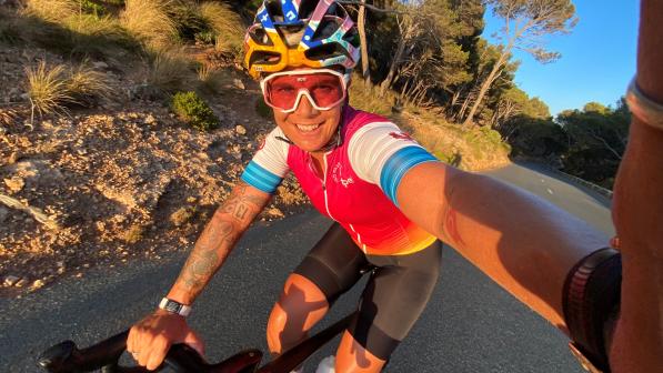 A woman is taking a selfie as she rides along a tarmac road in Mallorca. It’s sunset. She’s wearing cycling kit, including helmet and sunglasses. She’s smiling into the camera