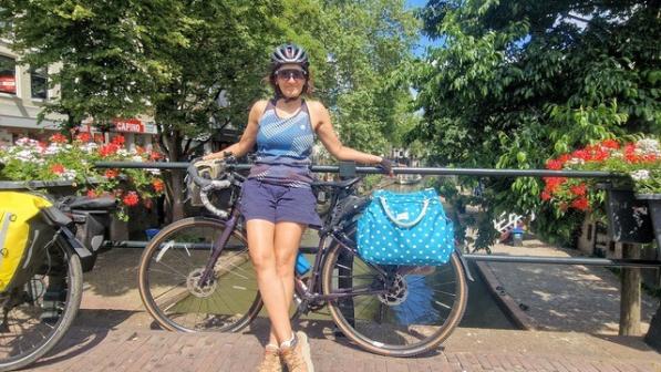 A woman is leaning against a touring bike leaning against a fence on a bridge across a canal. She is wearing shorts and a sleeveless T-shirt, sunglasses and a helmet. There are a lot of trees and flowers in planters in the background.