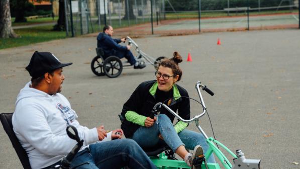 A man and a woman are on adapted cycles in a sports ground. They are both wearing jeans and fleeces