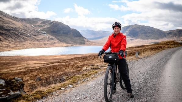 A woman is standing astride a packed off-road bike on a gravel track through mountainous countryside with a lake in the background. She is wearing red, winter cycling jacket, winter gloves and trousers, as well as a helmet. She is smiling at the camera