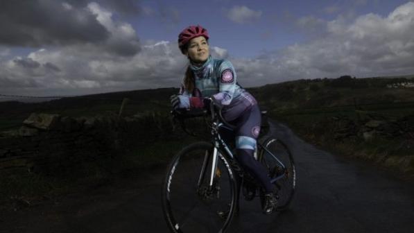Vicky Mathwin is standing astride her road bike with one foot on the pedal on a country lane. She is wearing full racing cycling kit and a helmet.