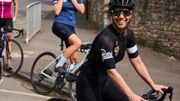 Rachael Burnside is sitting astride a red road bike. She is wearing black cycling shorts and a black and white short-sleeved jersey, as well as a helmet and cycling sunglasses. She’s smiling at the camera. In the background are two more women on road bikes.